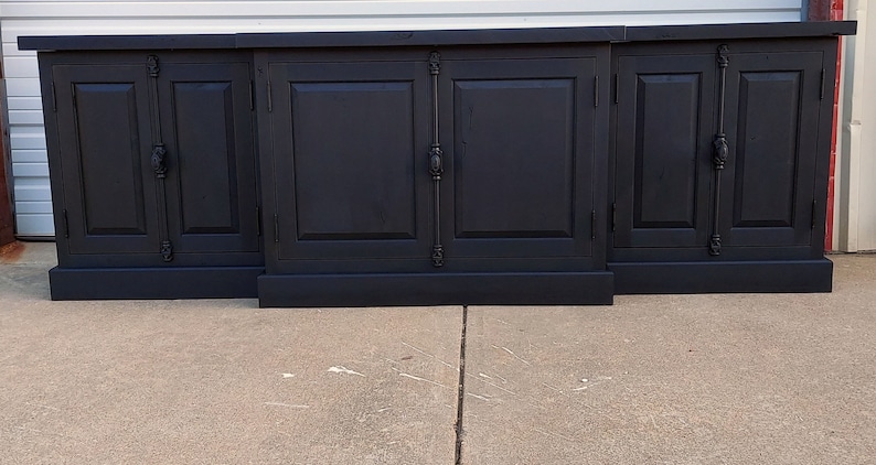May include: A black wooden cabinet with six doors and black metal hardware. The cabinet has a base and sits on a concrete floor.