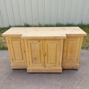 May include: A light wood cabinet with three doors and gold hardware. The cabinet has a simple design with a flat top and a slightly raised base.