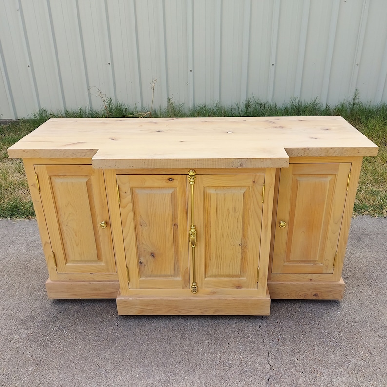 May include: A light wood cabinet with three doors and gold hardware. The cabinet has a simple design with a flat top and a slightly raised base.