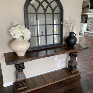 May include: A dark brown wooden console table with a lower shelf. The table has two turned legs with a decorative design. The table is decorated with a white vase with white flowers and a black vase with dried branches. A black arched mirror with a grid pattern is hanging on the wall behind the table.