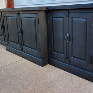 May include: A black wooden cabinet with four doors and ornate metal hardware. The cabinet has a dark wood finish and a simple design.