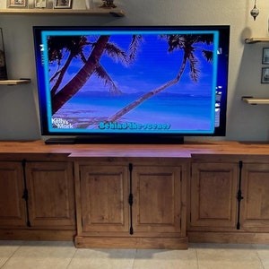 May include: A wooden entertainment unit with three cabinet doors. The cabinet is a dark brown colour and has black metal hardware. A flat screen TV is mounted above the cabinet. The TV screen shows a tropical beach scene with palm trees and the text "Behind the Scenes".