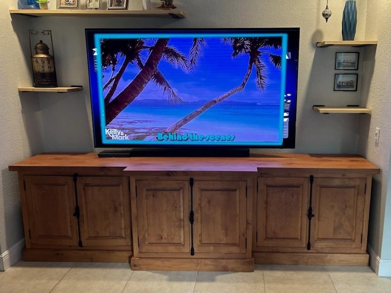 May include: A wooden entertainment unit with three cabinet doors. The cabinet is a dark brown colour and has black metal hardware. A flat screen TV is mounted above the cabinet. The TV screen shows a tropical beach scene with palm trees and the text "Behind the Scenes".