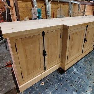 May include: A wooden cabinet with four doors and black metal hardware. The cabinet is made of light wood and has a natural finish. The cabinet is sitting on a black surface with a pattern of holes.