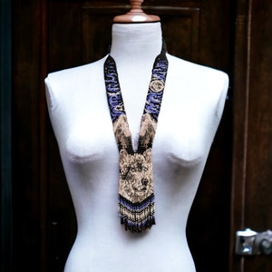 Könnte beinhalten: Eine Perlenkette mit einem Wolfsmuster in Braun-, Schwarz- und Lilatönen. Die Kette hat unten eine Fransenborte.