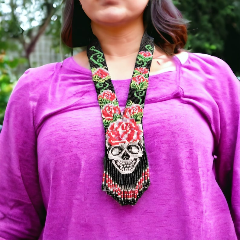 May include: A black beaded necklace with a skull and roses design. The skull is surrounded by red roses and green leaves. The necklace is long and hangs down to the chest.