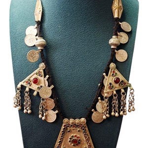 May include: An ornate necklace with a dark cord, featuring silver-toned pendants, coins, and triangular elements adorned with red and green gemstones. The necklace has a large central pendant with dangling chains.