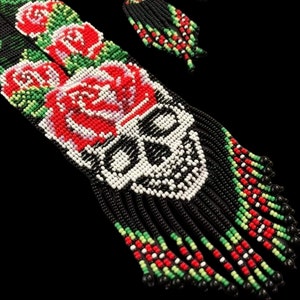 May include: A pair of beaded earrings with a skull and roses design. The earrings are black with red, white, and green beads. The skull is white with black details. The roses are red with white and green details.