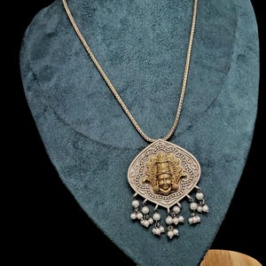 Pode incluir: Um colar com pingente prateado com uma figura de divindade dourada no centro. O pingente tem um design detalhado e é adornado com pequenas contas brancas peroladas. O colar é exibido em um busto verde-azulado.