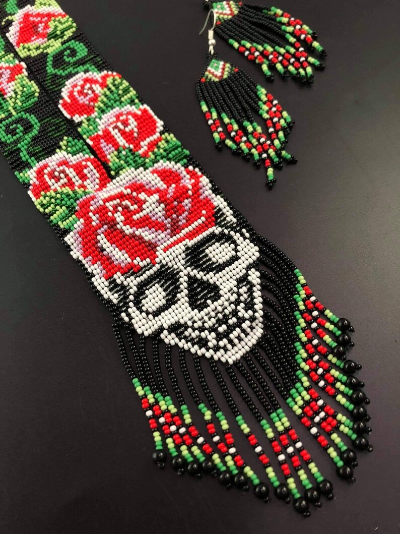 May include: A beaded necklace and earrings set with a skull design and red roses. The necklace has black, white, red, and green beads. The earrings are black, white, red, and green with black tassels.
