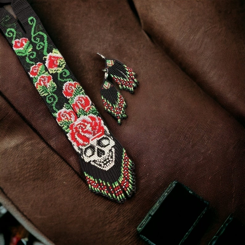 May include: A black beaded necklace with a skull and red roses design. The necklace is on a brown fabric background. Two matching beaded earrings are also pictured.