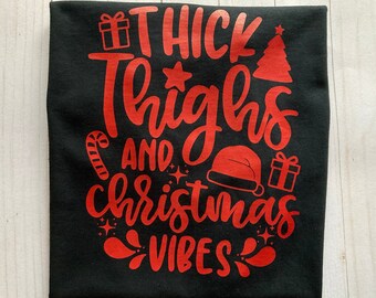 May include: Black t-shirt with red text that reads "Thick Thighs and Christmas Vibes". The text is surrounded by Christmas-themed icons like a gift box, candy cane, and a Christmas tree.