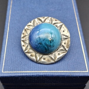 May include: A round, silver-toned brooch with a central, glossy blue orb. The orb is surrounded by a silver frame with a geometric design. The brooch is set against a blue background.