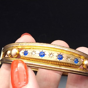 May include: A gold-toned hinged bangle bracelet featuring a central horizontal bar with three round blue stones and three small clear stones set in a star pattern. The bracelet has smooth gold spheres on each side of the central decorative bar.