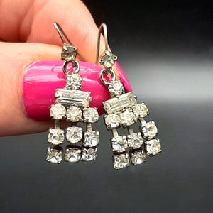 Può includere: Orecchini a lampadario color argento con strass trasparenti e scintillanti. Ogni orecchino presenta una parte superiore rettangolare, una fila di pietre rettangolari e tre file di pietre rotonde, creando un effetto a cascata. Chiusura a gancio.