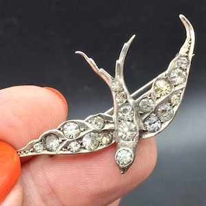 May include: A silver-toned swallow bird brooch featuring a detailed metal body encrusted with numerous clear, sparkling rhinestones. The bird is depicted in flight with wings spread wide, showcasing a textured metallic finish and a pin mechanism.