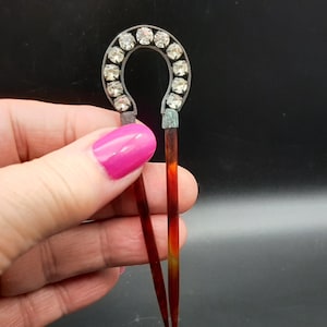 Puede incluir: Una horquilla vintage con una parte superior en forma de herradura adornada con pedrería transparente. La base del pasador es de color marrón rojizo oscuro. El accesorio para el cabello se sostiene sobre un fondo oscuro.