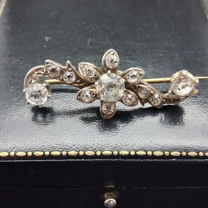 Op de afbeelding: Een zilverkleurige metalen broche met een centraal bloemvormig cluster van heldere, gefacetteerde stenen. Het ontwerp loopt uit in een gebogen, rankachtige vorm versierd met extra heldere stenen, alles geplaatst op een metalen basis.