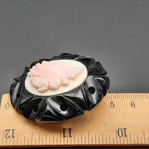 May include: A vintage oval brooch featuring a cameo of a woman's profile in pink and white. The cameo is set in a black, carved, floral-shaped frame. The brooch measures approximately 2.5 inches in length.
