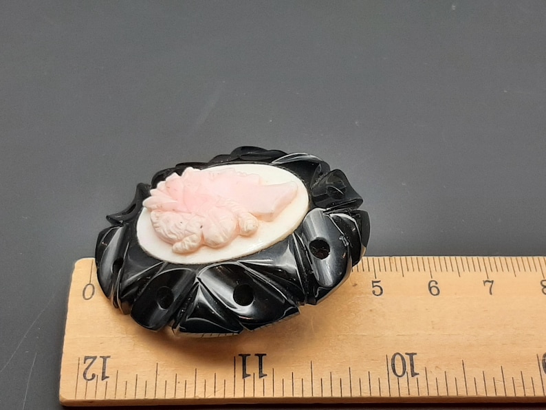 May include: A vintage oval brooch featuring a cameo of a woman's profile in pink and white. The cameo is set in a black, carved, floral-shaped frame. The brooch measures approximately 2.5 inches in length.