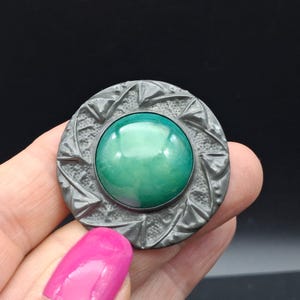 May include: A circular brooch featuring a smooth, polished, dome-shaped green stone set in the center. The surrounding metal frame is dark gray with a textured, rugged surface and a repeating pattern of raised triangular shapes along the edge.