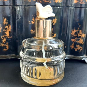 May include: A clear glass bottle with a gold top and a white bird-shaped stopper. The bottle has a textured design and is filled with a clear liquid.