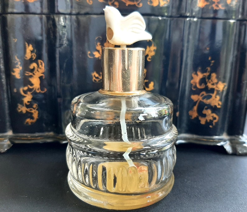 May include: A clear glass bottle with a gold top and a white bird-shaped stopper. The bottle has a textured design and is filled with a clear liquid.