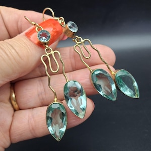 May include: A pair of gold-toned dangle earrings featuring a small round light blue gemstone at the top, connected by a wavy metal wire to two larger, teardrop-shaped light blue gemstones hanging at the base of each earring.