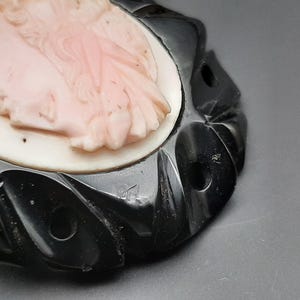 May include: A vintage cameo brooch featuring a carved profile of a woman in a light pink hue. The cameo is set against a white background and framed by a black, intricately designed border. The brooch has a classic, elegant aesthetic.