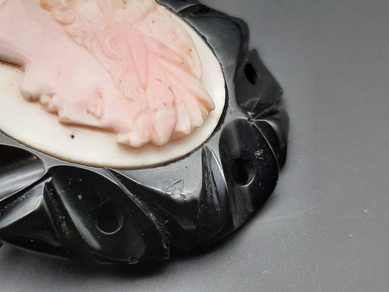 May include: A vintage cameo brooch featuring a carved profile of a woman in a light pink hue. The cameo is set against a white background and framed by a black, intricately designed border. The brooch has a classic, elegant aesthetic.