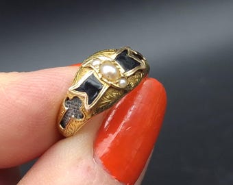 Può includere: Un anello in oro con un intricato motivo inciso, accenti in smalto nero e due piccole perle bianche incastonate al centro. La fascia presenta un motivo decorativo con un intaglio a forma di croce e una finitura lucida con dettagli in metallo.