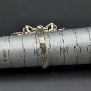 May include: Close-up of a silver ring with a decorative design, resting on a metal ring sizer. The ring has a detailed pattern and is positioned to show its profile. The ring sizer has measurement markings.