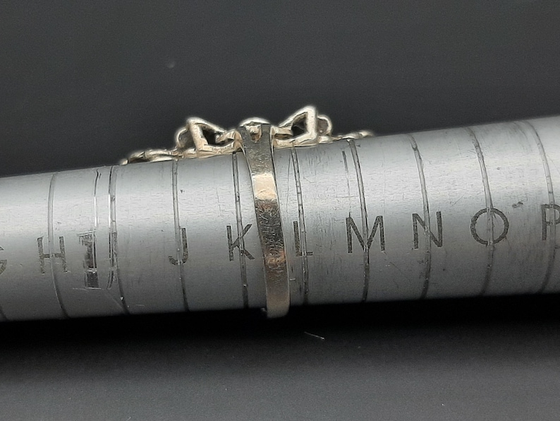 May include: Close-up of a silver ring with a decorative design, resting on a metal ring sizer. The ring has a detailed pattern and is positioned to show its profile. The ring sizer has measurement markings.
