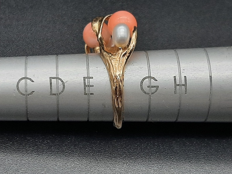 Pu&ograve; includere: Anello in oro con tre pietre di corallo e perla. L'anello &egrave; posto su un righello di metallo con segni di misurazione.