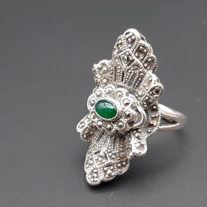 May include: A silver-toned ring with a large, ornate design. The ring features a central oval-shaped, green gemstone, surrounded by a detailed, floral-like pattern. The band is split at the top, adding to the ring's unique style.