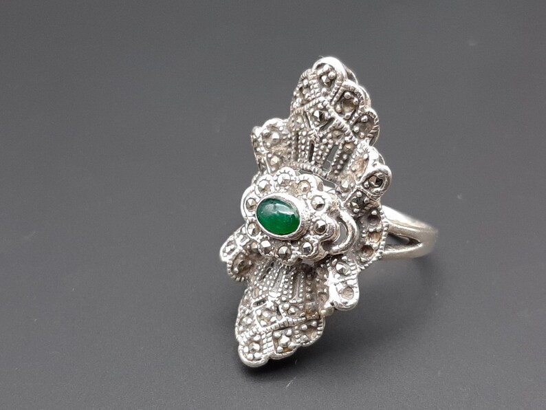 May include: A silver-toned ring with a large, ornate design. The ring features a central oval-shaped, green gemstone, surrounded by a detailed, floral-like pattern. The band is split at the top, adding to the ring's unique style.