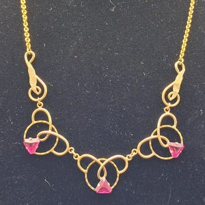 May include: A gold-tone necklace featuring a delicate chain and three symmetrical, swirling metal links. Each link holds a small, triangular, vibrant pink gemstone. The design incorporates open, curved metalwork that connects the stones.