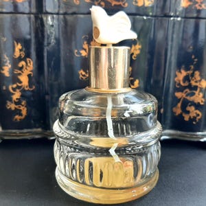 May include: A clear glass bottle with a gold top and a white ceramic stopper. The bottle has a textured design and is filled with a clear liquid.