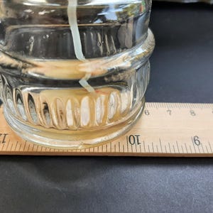 May include: A clear glass jar with a textured surface sits on a wooden ruler marked with inches. The jar contains a white candle with a yellow wax pool at the bottom.