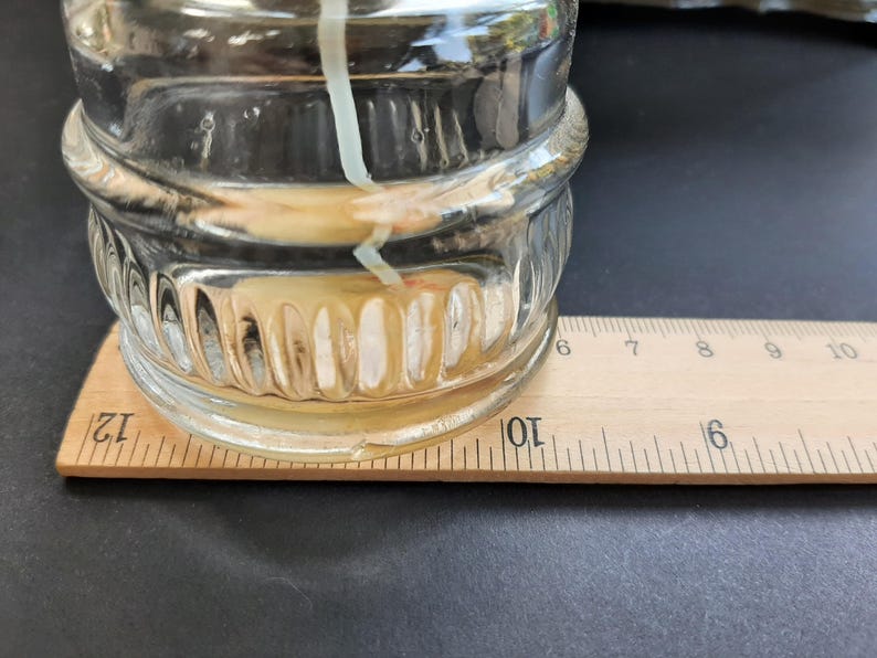 May include: A clear glass jar with a textured surface sits on a wooden ruler marked with inches. The jar contains a white candle with a yellow wax pool at the bottom.