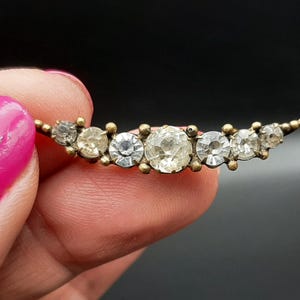 Può includere: Spilla vintage con design curvo, incastonata con sette strass rotondi e trasparenti in metallo dorato. Gli strass variano di dimensioni e brillano. Un classico gioiello.