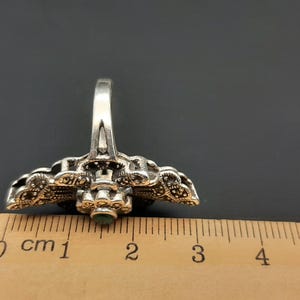 May include: A silver-toned ring with an ornate design, featuring small, dark stones and a central green stone. The ring is placed next to a ruler, showing its size.