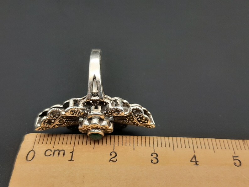 May include: A silver-toned ring with an ornate design, featuring small, dark stones and a central green stone. The ring is placed next to a ruler, showing its size.