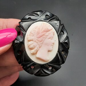 May include: Oval cameo brooch featuring a profile of a woman in a light pink hue, set against a white background. The cameo is framed by a black, ornate, openwork border. A pin is attached to the back.