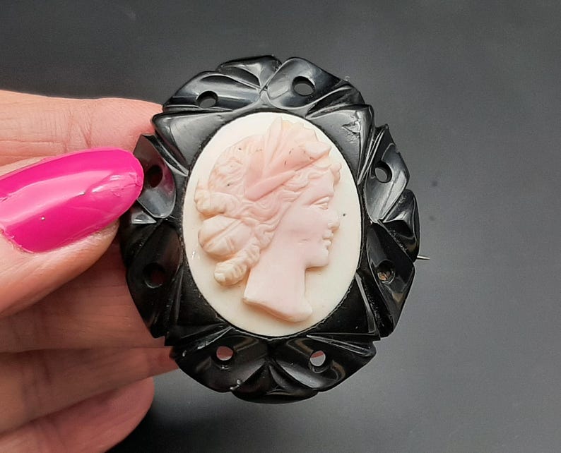 May include: Oval cameo brooch featuring a profile of a woman in a light pink hue, set against a white background. The cameo is framed by a black, ornate, openwork border. A pin is attached to the back.