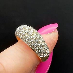 May include: A gold-toned band ring featuring a wide, domed top surface densely encrusted with multiple small, clear, round-cut stones. The metal band has a smooth, polished finish on the sides and bottom, contrasting with the textured, sparkling stone-set top.