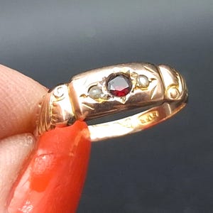 以下が含まれることがあります： 中央に丸いファセットカットの深い赤色のガーネットを配し、両脇に小さな白い真珠をあしらったゴールドリング。バンドには彫刻が施された長方形のセッティングがあり、側面には装飾的なスクロール模様、内側には刻印が見える。