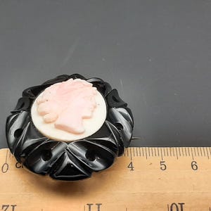 May include: A vintage black and pink cameo brooch. The brooch features a pink profile set against a white background, surrounded by a black, ornate frame. The brooch is approximately 2 inches in diameter.