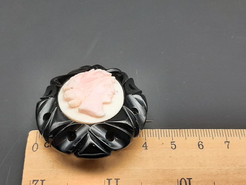 May include: A vintage black and pink cameo brooch. The brooch features a pink profile set against a white background, surrounded by a black, ornate frame. The brooch is approximately 2 inches in diameter.