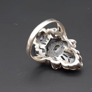 May include: Silver-toned ring with a floral design. The ring features a central dark stone surrounded by intricate metalwork. The band is smooth and rounded.